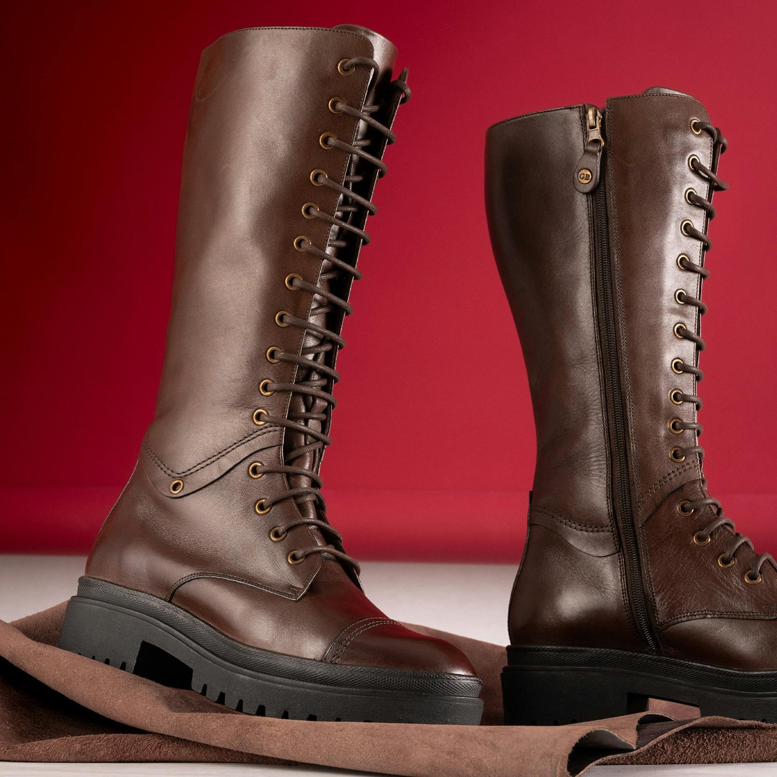 Elegant brown leather knee-high boots with lace and zipper on red background.